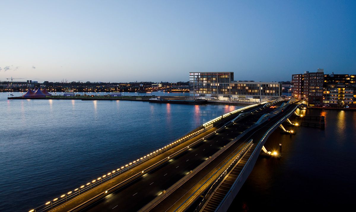 Dusk view across the water to a city waterfront, a lit road curving along the shore — urban visualisation
